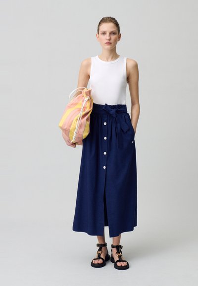 Jupe midi navy avec des boutons sur le devant et une ceinture à nouer, associée à un haut sans manches blanc et un sac à cordon multicolore. Sandales noires.