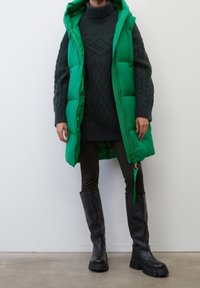 Grøn puffervest med hætte, der har en frontlynlås og quiltet tekstur. Båret over en mørk striktrøje og mørke bukser, parret med knæhøje sorte støvler.