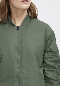 Giacca bomber verde con zip frontale, colletto a coste e tessuto trapuntato. Presenta tasche laterali e elementi di design snelli.