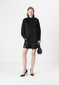 Camisa com franjas pretas e design com colarinho, combinada com calções de pele pretos brilhantes. Modelo segura uma pequena mala preta, usando tacões pretos.
