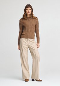 Suéter de punto trenzado marrón combinado con pantalones de pierna ancha beige y botines marrones. El suéter presenta un escote redondo y puños acanalados.