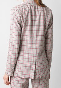 Bagview af en person iført en hvid blazer og bukser med røde og sorte tern, som står foran en enkel lys baggrund.