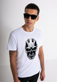 Camiseta blanca de algodón con un gran gráfico de calavera en negro y plateado con ojos verdes. El modelo lleva gafas de sol negras.