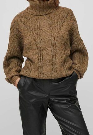 Pull à col roulé en maille torsadée marron épais, associé à un pantalon en cuir noir taille haute, les mains posées négligemment dans les poches.