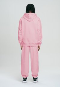 Růžová oversized mikina a odpovídající tepláky vyrobené z měkké látky, se žebrovanými manžetami a kapucí se šňůrkou, doplněné černými teniskami.