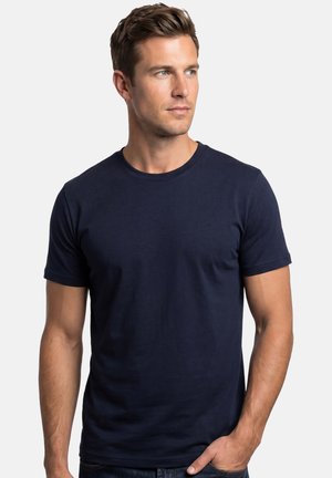 Junger Mann mit kurzem braunem Haar, der ein schlichtes dunkelblaues T-Shirt mit kurzen Ärmeln trägt und nach rechts schaut, mit einer Hand in der Tasche.