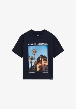 Tengerészkék póló pálmafák és napfényes épület grafikájával, "Puerto Montaña" felirattal, és a kép alatt földrajzi koordinátákkal.