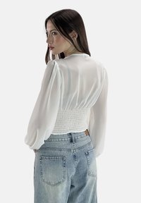Blusa bianca con maniche lunghe trasparenti e schiena arricciata. Abbinata a jeans azzurri a vita alta. Tessuto morbido, design minimal.