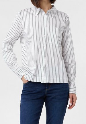 Femme portant une chemise blanche à manches longues avec de fines rayures verticales noires et un jean bleu foncé, debout avec une main dans la poche.
