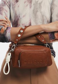 Borsa a tracolla in pelle marrone con logo "Desigual" impresso, dotata di una tracolla rimovibile con borchie argentate, chiusura con zip e superficie texturizzata.