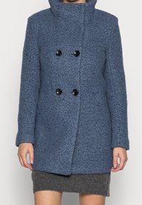 Modrý plášť z vlněné směsi s texturovaným povrchem, dvojřadý střih, velké černé knoflíky a vysoký límec. Dlouhé rukávy a střih sahající k bokům.