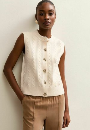 Cream sleeveless knit vest with cable pattern, featuring gold-tone floral buttons and a round neckline, worn with beige satin trousers.