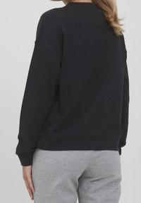 Felpa nera con una vestibilità morbida, scollatura rotonda e fondo a costine. Abbinata a pantaloni della tuta grigi, che mostrano uno stile casual e confortevole.