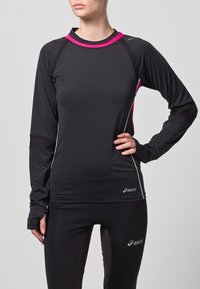 Donna che indossa un top sportivo a maniche lunghe nero e rosa e leggings neri, entrambi con il logo Asics, mentre posa con una mano sui fianchi.
