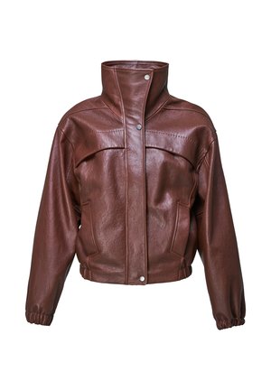 Femme portant une veste en cuir marron, un pantalon large gris, tenant un sac à main marron, et des talons pointus noirs sur un fond gris uni.