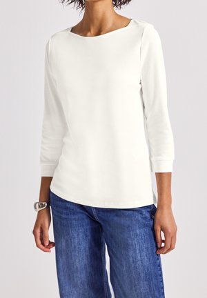 Mujer con una camiseta blanca de manga larga lisa y vaqueros azules, de pie con las manos relajadas a los lados contra un fondo neutro.