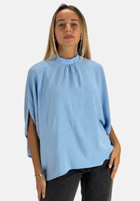 Lyseblå oversized top med høj hals, struktureret stof og korte ærmer. Kombineret med mørke jeans, hvilket skaber en løs pasform.