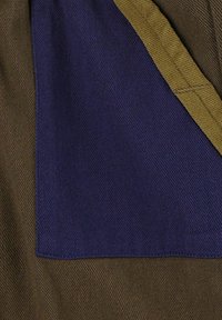 Close-up van stof met gelaagde bruine, marineblauwe en mosterdkleurige textielstukken die diagonale weefpatronen en stikdetails tonen.