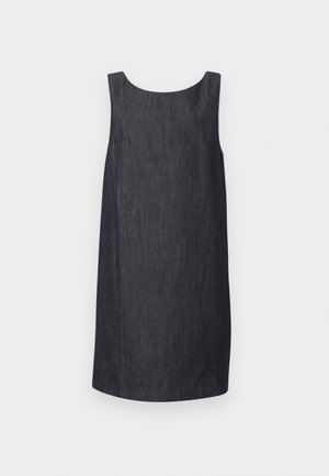 Jeune femme aux longs cheveux noirs portant une robe sans manches gris foncé jusqu'aux genoux et des sandales noires à brides, debout devant un fond gris clair.