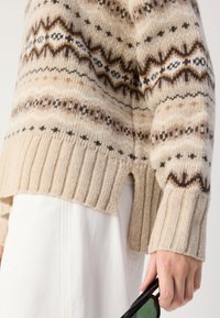 Pull en tricot beige et marron avec des motifs géométriques. Doté d'un ourlet côtelé et d'évasements sur les côtés. Le modèle tient des lunettes de soleil dans une main.