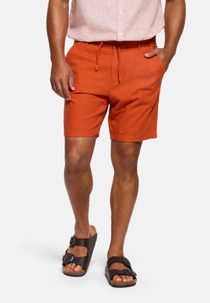 Hombre con pantalones cortos naranjas con cordón, camisa de manga corta rosa claro y sandalias marrones oscuras de doble correa, de pie con las manos en los bolsillos.