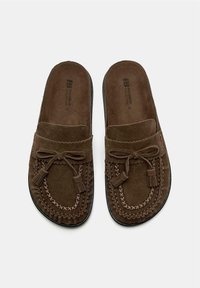 Chaussons mocassins en daim marron à enfiler avec des coutures décoratives et des détails de nœud à frange sur fond blanc.