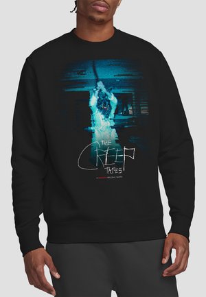 Person wearing black sweatshirt featuring distorted blue figure with a mask holding a rope, text "The Creep Tapes" on front center.