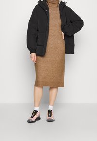 Doudoune noire à capuche, robe en maille beige, et chaussures de sport colorées à semelle texturée, portées avec des chaussettes blanches.