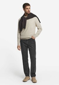 Beige gestrickter Pullover mit rundem Ausschnitt, dunkelgraue Jeans und hellbraune Schuhe. Ein dunkler Schal ist über die Schultern gelegt.