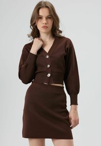 Bruine, getextureerde, cropped cardigan met een V-hals en drie zilveren knopen, gecombineerd met een bijpassende bruine rok.
