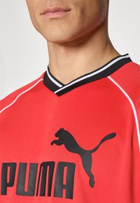 Rød sportsdrakt med svart V-hals, med en fremtredende svart Puma-logo og hvit kanting langs skuldrene.