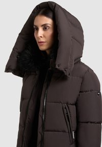 Jachetă impermeabilă maro, căptușită, cu un glugă mare, margine din blană sintetică, închidere cu fermoar pe față și buzunare laterale. Textură matlasată cu un finisaj neted.