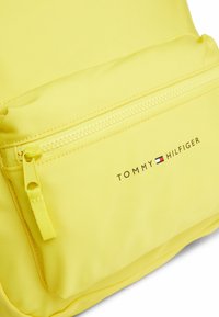Mochila amarilla con un tejido texturizado, que presenta un bolsillo frontal con cremallera y el logo "TOMMY HILFIGER" impreso en negro.