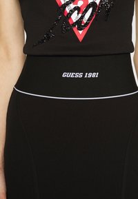 Czarna spódnica typu wysoki stan z białym logo "GUESS 1981" na pasku, zestawiona z czarnym bezrękawnikiem z czerwono-czarnym zdobieniem z cekinów.
