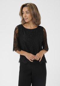 Top in pizzo nero con maniche in chiffon a tre quarti e orlo a scallop. Motivi floreali intricati su tutta la superficie, abbinato a pantaloni neri.