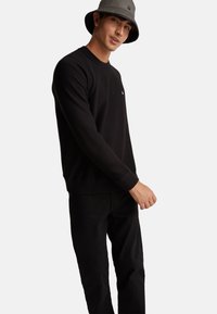 Maglione nero lavorato a maglia con polsini e orlo a coste, abbinato a jeans neri. Un cappello a secchiello grigio presenta un piccolo logo decorativo.