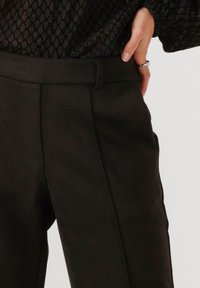 Zwarte, op maat gemaakte broeken met een gladde textuur, voorzien van een hoge taille, voorplooien en zijzakken. Draagt een patroonrijke, doorzichtige top.