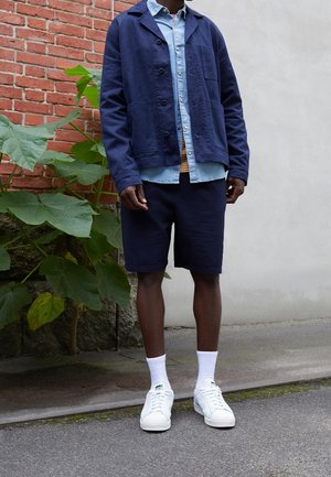 Homme portant une veste bleu marine, une chemise bleu clair, un short bleu marine, des chaussettes blanches et des baskets blanches, debout près de murs en briques et en béton avec des plantes vertes.