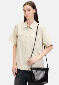 Beżowa koszula na krótki rękawek z zamkiem błyskawicznym i dwoma kieszeniami na piersi, zestawiona z czarną skórzaną torbą na ramię. Torba ma gładką teksturę i regulowany pasek.
