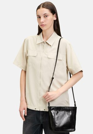 Beige Kurzarm-Reißverschlusshemd mit zwei Brusttaschen, kombiniert mit einer schwarzen Leder-Crossbody-Tasche. Die Tasche hat eine glatte Textur und einen verstellbaren Riemen.
