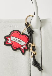 Balta tekstūruota rankinė su juoda oda ir auksine grandine, su raudona širdies formos etiketė, pažymėta „ALDO“, prikabinta prie rankenos.