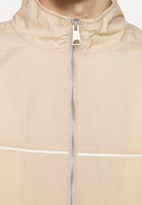 Beige jacka med uppfällt krage och silverfärgad dragkedja som har en liten Jumpman-logotyp på dragkedjehandtaget.