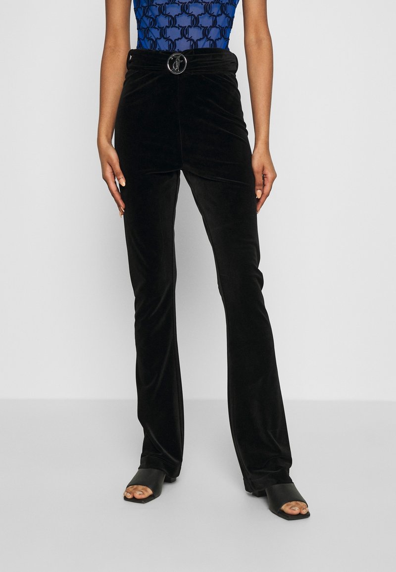 Femme portant un pantalon taille haute en velours noir avec une boucle de ceinture en anneau argenté et des talons ouverts noirs, se tenant devant un fond uni.