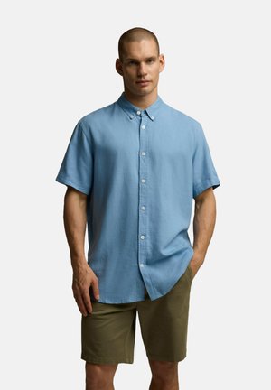 Jasnoniebieska koszula zapinana na guziki, wykonana z miękkiego materiału, z krótkimi rękawami, kołnierzykiem i luźnym krojem. W połączeniu z oliwkowymi szortami.
