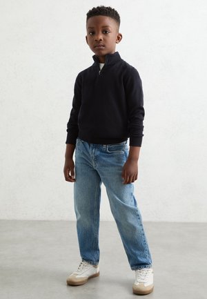 Junge steht und trägt einen schwarzen Pullover mit Reißverschluss vorne, hellblaue Jeans und weiße Turnschuhe vor einem einfarbigen hellen Hintergrund.