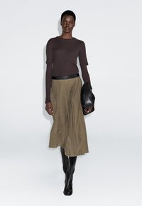 Femme portant un haut marron à manches longues, une jupe midi plissée olive, une ceinture noire, des bottes noires montantes, tenant un sac en cuir noir.