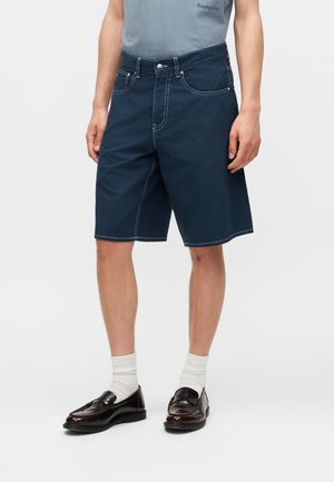 Ένας άνδρας φοράει μπλε σκούρες βερμούδες μέχρι τα γόνατα με λευκή ραφή, λευκές κάλτσες μέχρι τον αστράγαλο, σκουρόχρωμα καφέ δερμάτινα μοκασίνια και ένα ανοιχτό μπλε πουκάμισο.