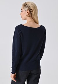 Marineblauer Pullover mit weitem Ausschnitt und langen Ärmeln, der Knopfdetails an den Bündchen aufweist. Weicher Stoff mit mittlerer Textur.