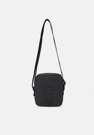 Kompaktowa czarna torba crossbody z regulowanym paskiem, z naszywką z logo Tommy Jeans z przodu, na jednolitym białym tle.