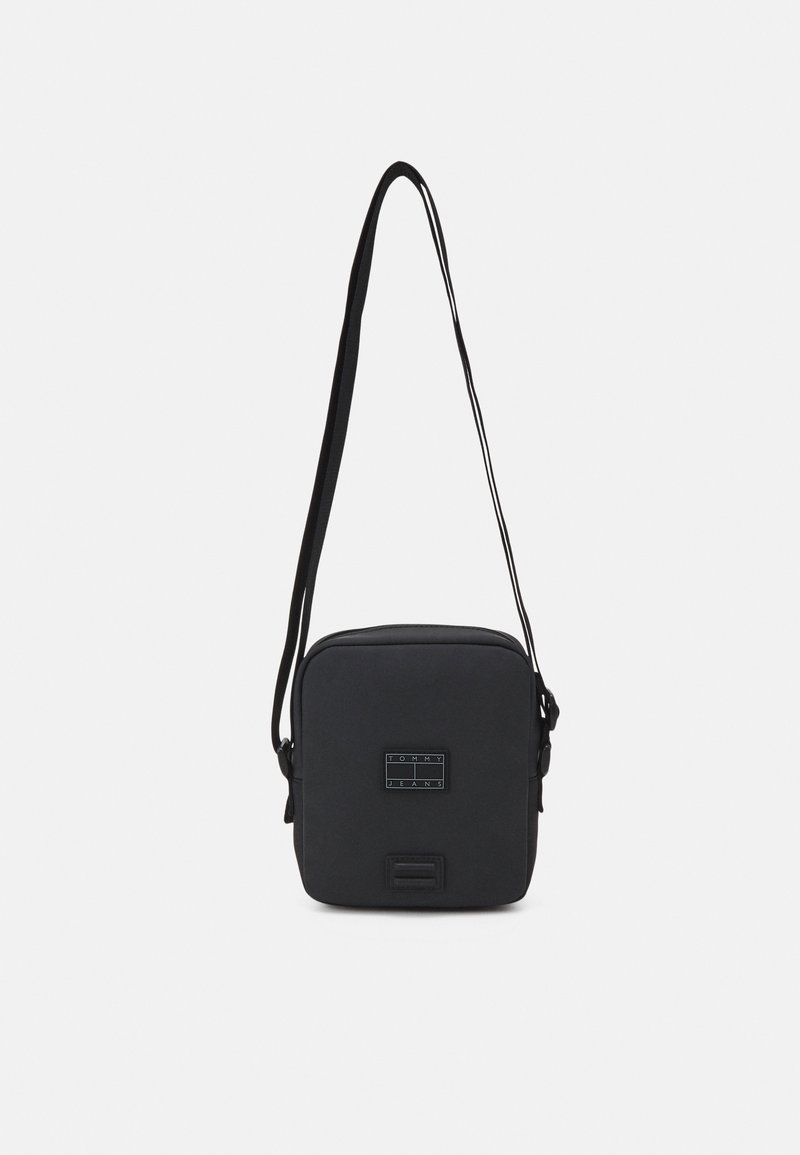 Borsa a tracolla compatta nera con tracolla regolabile, con patch del logo Tommy Jeans sul davanti, su uno sfondo bianco semplice.